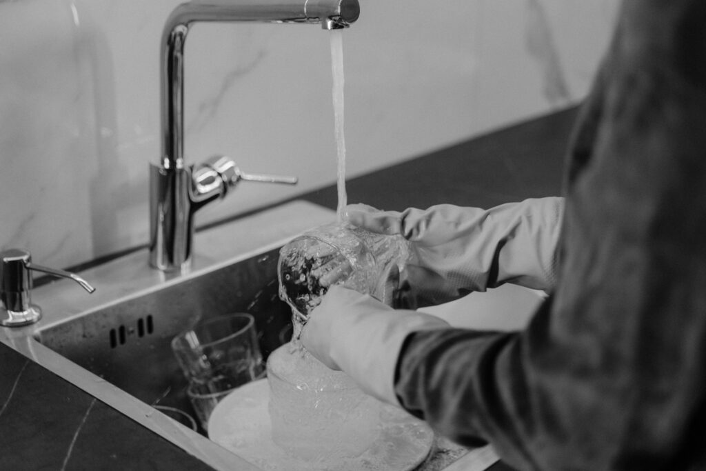 Monochrome photo of a person washing dishes under running water in a kitchen sink.